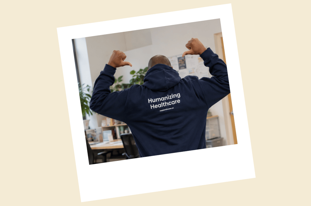 A person seen from behind in an office, pointing both thumbs at the words “Humanizing Healthcare” printed on the back of their navy hoodie.